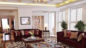 A spacious living room with a large red sofa, a beige armchair, and a coffee table with books and a vase. There are two lamps on side tables, and a large window with a view of the outside. - Travelers Safari - Westlands (Nairobi)