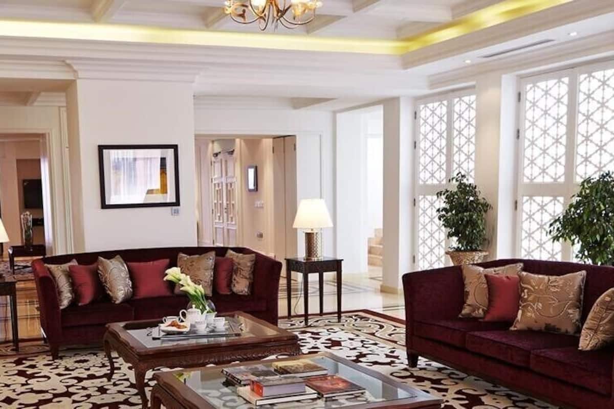 A spacious living room with a large red sofa, a beige armchair, and a coffee table with books and a vase. There are two lamps on side tables, and a large window with a view of the outside.