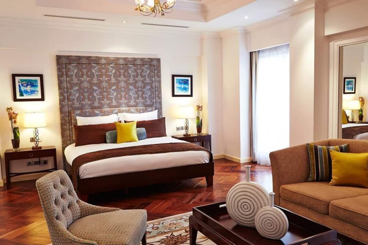 A hotel room with a large bed, a sofa, a chair, and a small table. There are two nightstands with lamps and a mirror.