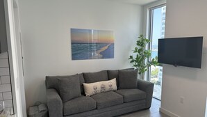 A modern living room with a grey sofa, a flat-screen TV mounted on the wall, a large window with a view of the sea, and a painting of a beach scene. - Premier Studio with King Bed and Queen Sofa-bed (Miami)