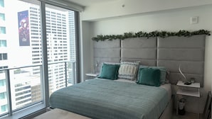 A modern bedroom with a large bed, a bench, and a view of tall buildings through a large window. - Premier Studio with King Bed and Queen Sofa-bed (Miami)
