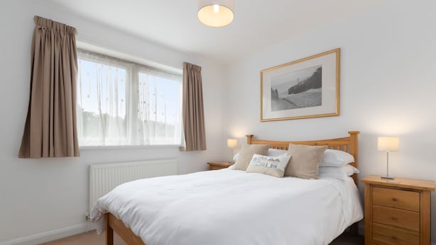 Une chambre à coucher comprenant un lit, des tables de chevet, une commode, une fenêtre avec des rideaux, un radiateur et un tableau encadré accroché au mur.