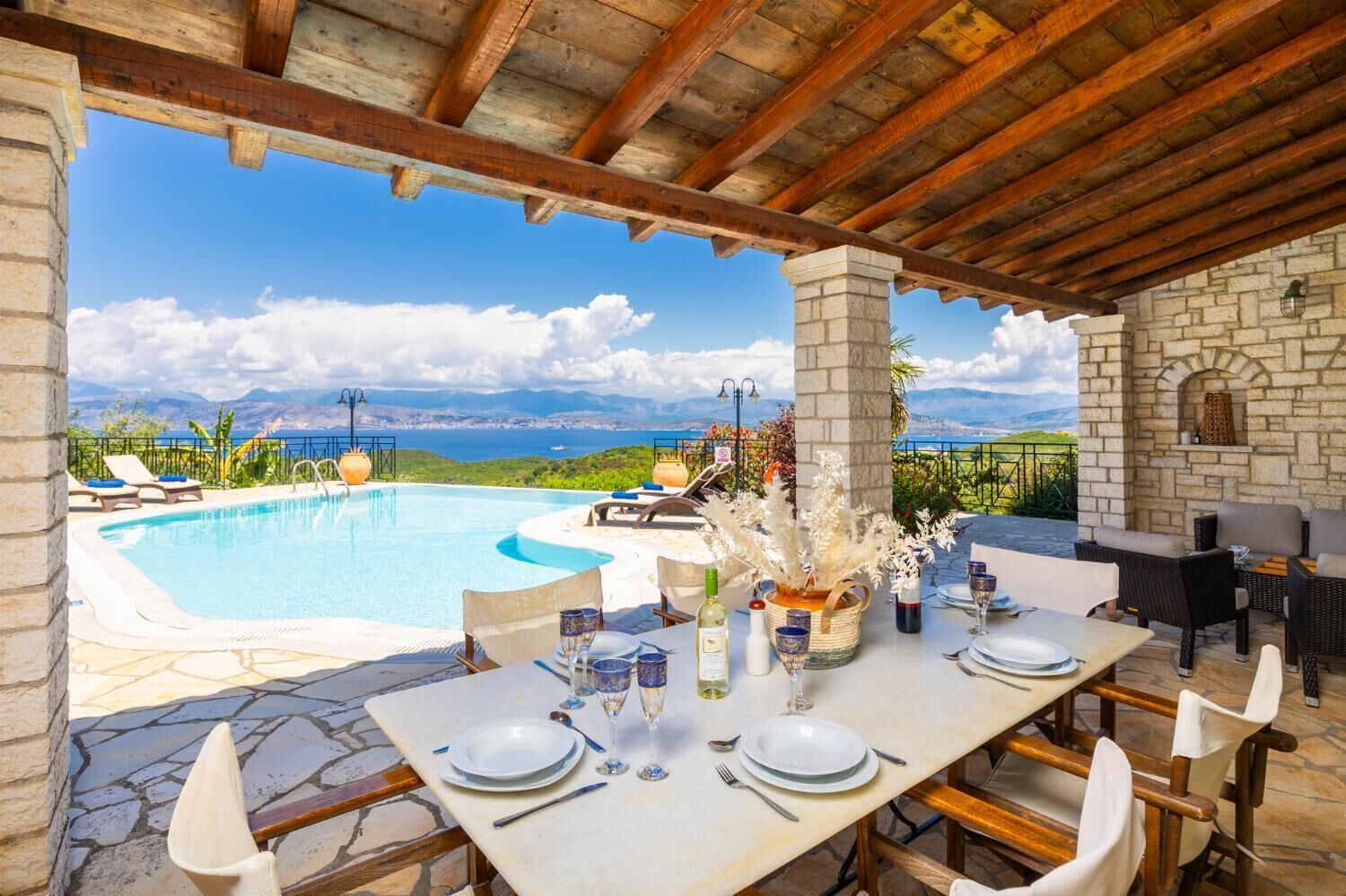 A poolside dining area with a view of the sea and mountains, stone pillars, and outdoor furniture.
