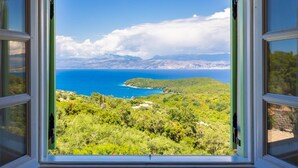 Blick aus dem Fenster auf eine Küstenlandschaft mit blauem Meer, grünen Hügeln und fernen Bergen.