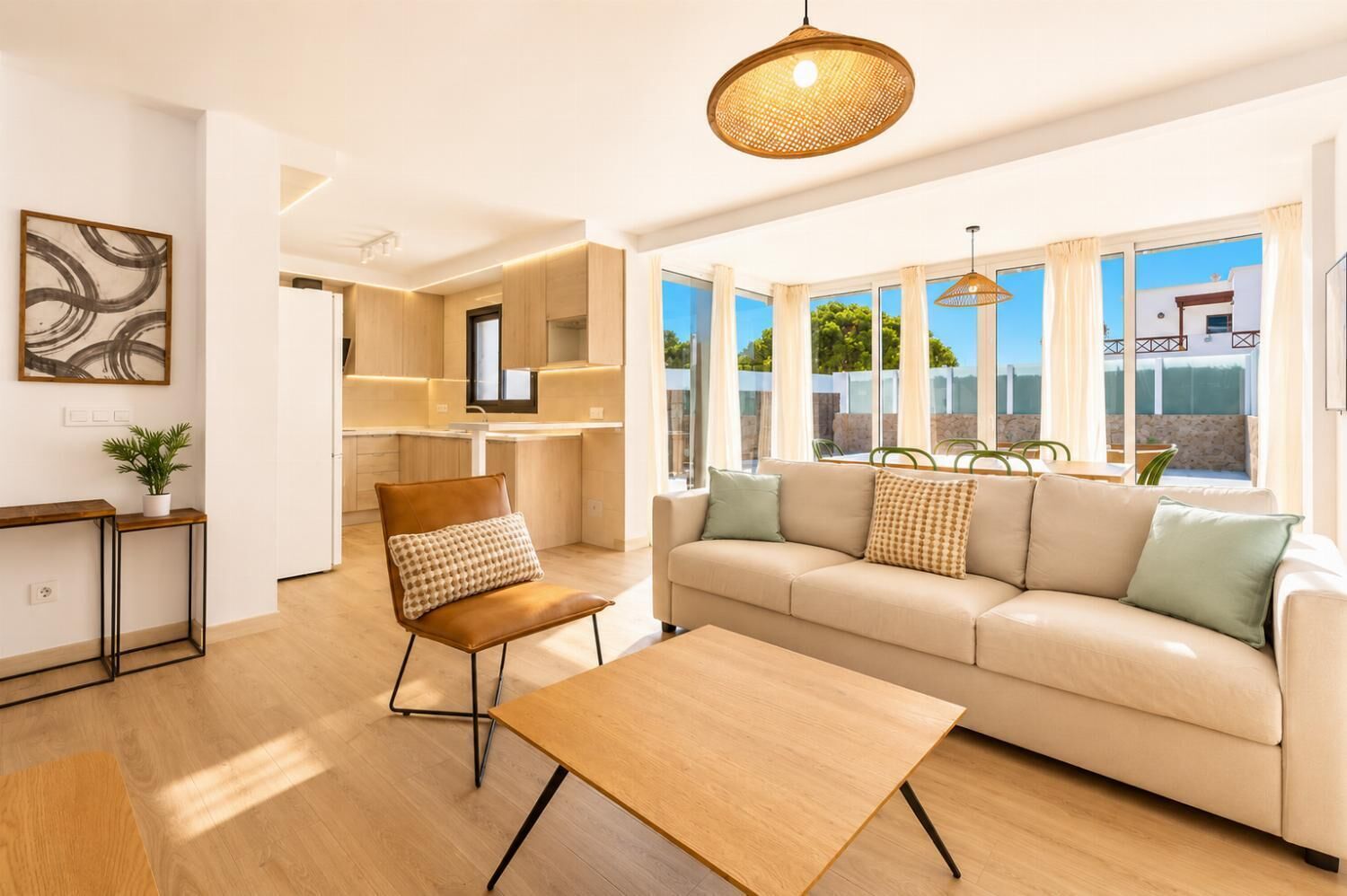 Un salon moderne avec un canapé, une table basse et un fauteuil. On aperçoit un coin repas avec une table et des chaises à travers de grandes fenêtres.