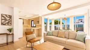 Un salon moderne avec un canapé, une table basse et un fauteuil. On aperçoit un coin repas avec une table et des chaises à travers de grandes fenêtres.