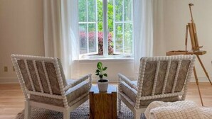 A room with two upholstered chairs facing a large arched window with white curtains, a small wooden table with a potted plant, and a painting on a tripod stand.