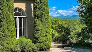 A house with a large arched window, surrounded by tall, green trees and a gravel driveway leading to a stone pathway.