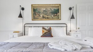 A neatly made bed with white linens, a colorful pillow, and a folded white towel on the bed. There are two bedside tables with lamps and a painting above the bed. - Cozy, Bright Suite w/ King Bed (Travelers Rest)
