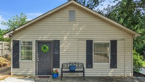 A single-story house with a front door, two windows with shutters, a small porch with a bench, and a wreath on the door. - Cozy, Bright Suite w/ King Bed (Travelers Rest)