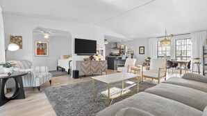 Moderni olohuone, jossa on sohva, sohvapöytä ja tuoleja. Taustalla näkyy keittiö ja seinälle on asennettu litteänäyttöinen televisio.