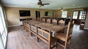 Un vaste espace dédié à la salle à manger, avec une longue table en bois et des chaises assorties, un ventilateur de plafond et un téléviseur fixé au mur.