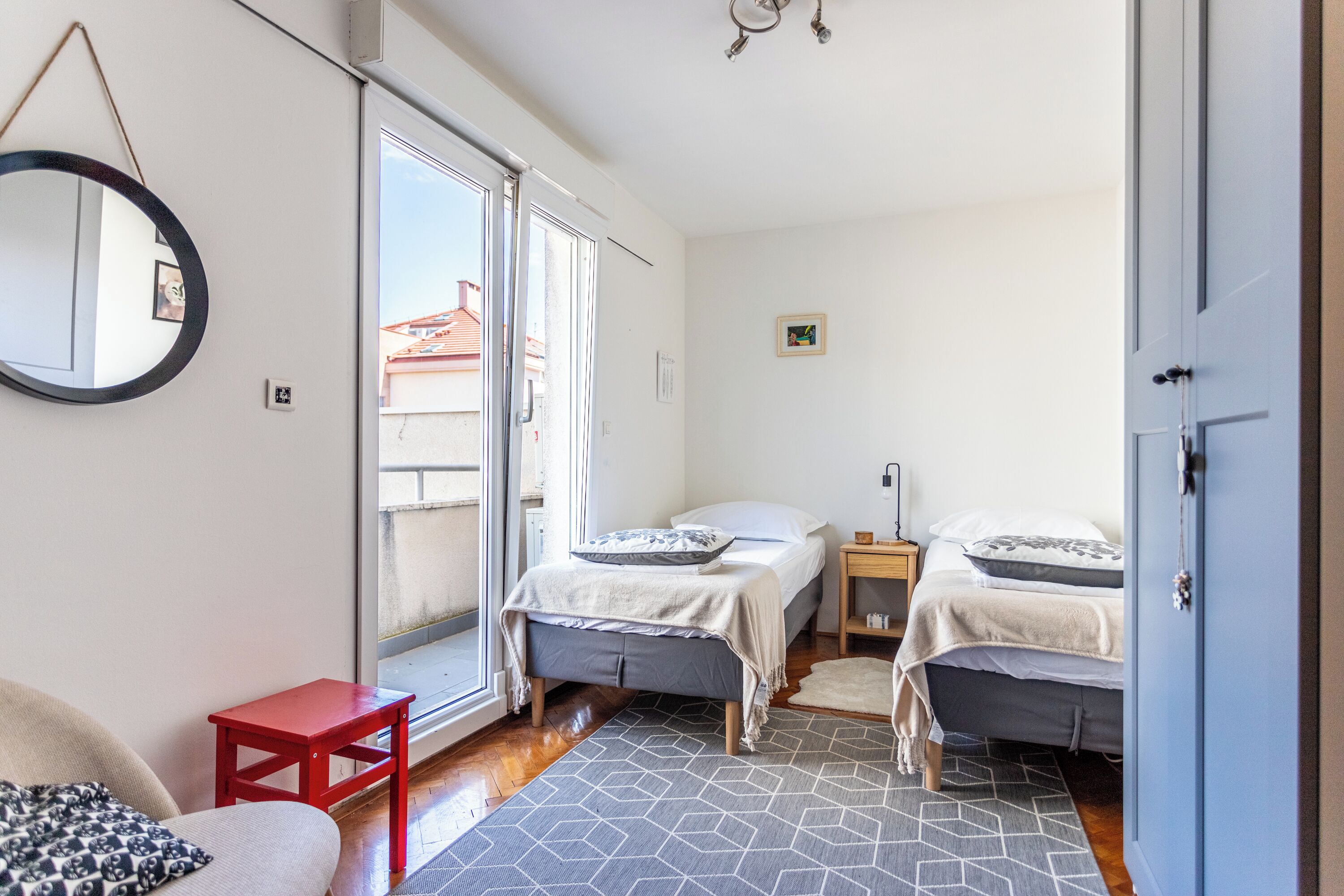A bedroom with two beds, a small table, a red stool, a mirror, and a view of the outside.
