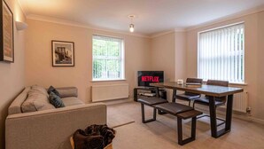A living room with a sofa, a coffee table, a TV displaying 'NETFLIX', and two windows with blinds. - Closetocitycentre&nec-business&family-freeparking (Solihull)