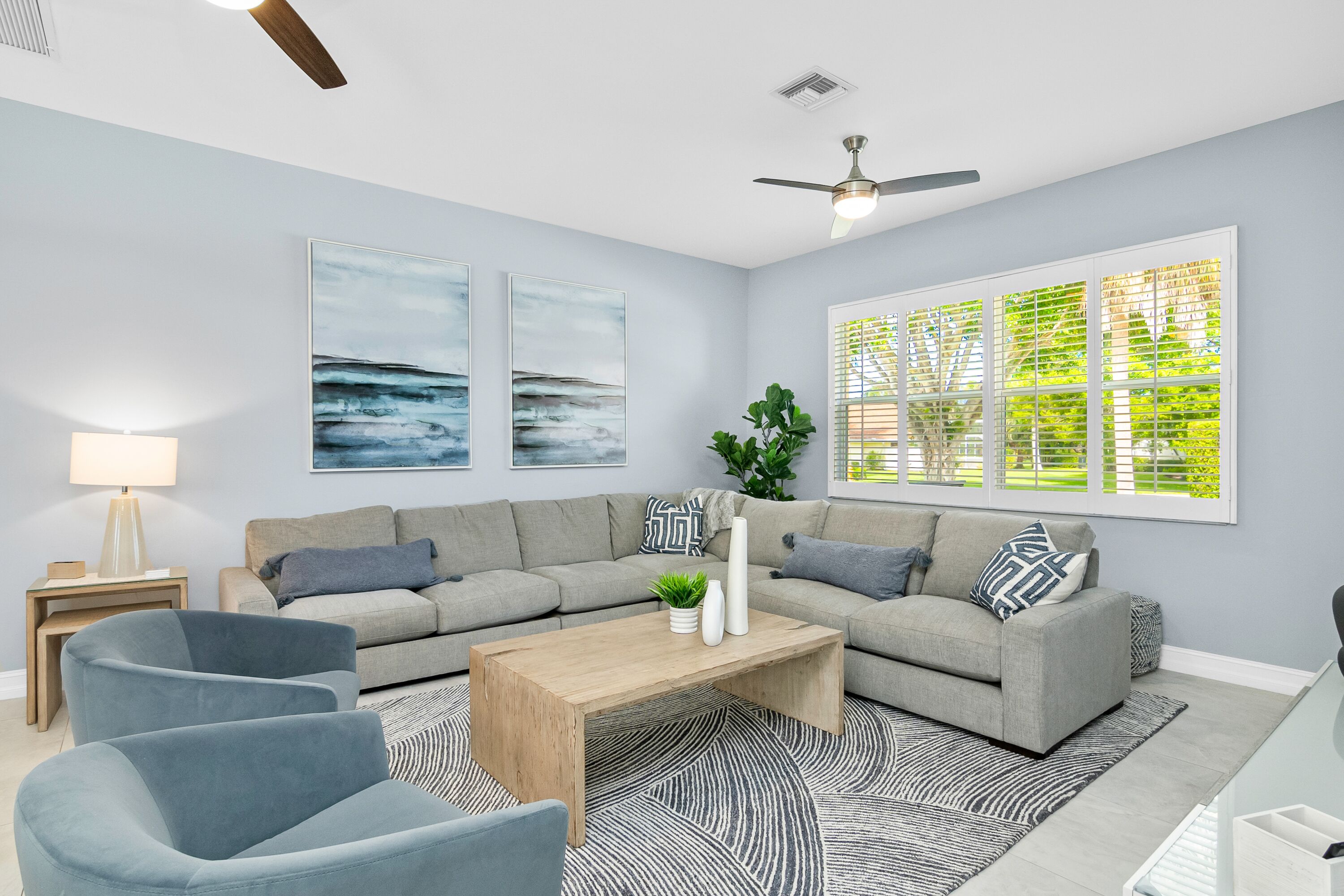Ein modernes Wohnzimmer mit einem grauen Sofa, zwei Sesseln, einem Holztisch und einem gemusterten Teppich. An den Wänden hängen zwei Bilder, eine Stehlampe und ein Fenster mit Jalousien.
