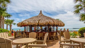 An outdoor bar area with wooden chairs and tables, a thatched roof, and a view of the ocean.