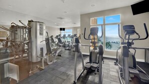 A gym with various exercise machines, a large window with a view of the ocean, and a flat-screen TV mounted on the wall.