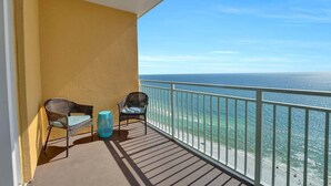 A balcony with two wicker chairs and a small blue stool, overlooking a beach with a clear view of the ocean.