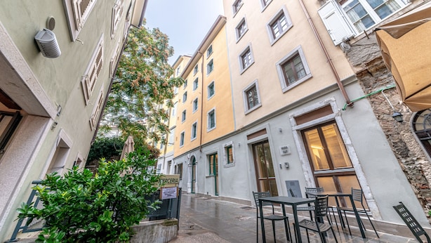 A narrow street with buildings on both sides, outdoor seating area with tables and chairs, and a signboard. - Hotel Mura 5&7 (Trieste)