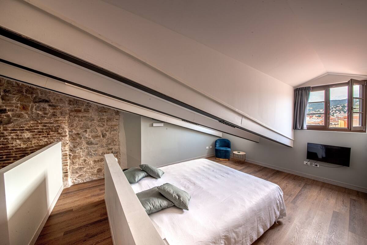 A bedroom with a sloped ceiling, a stone wall, a bed with pillows, a blue chair, a TV, and a window with a view.