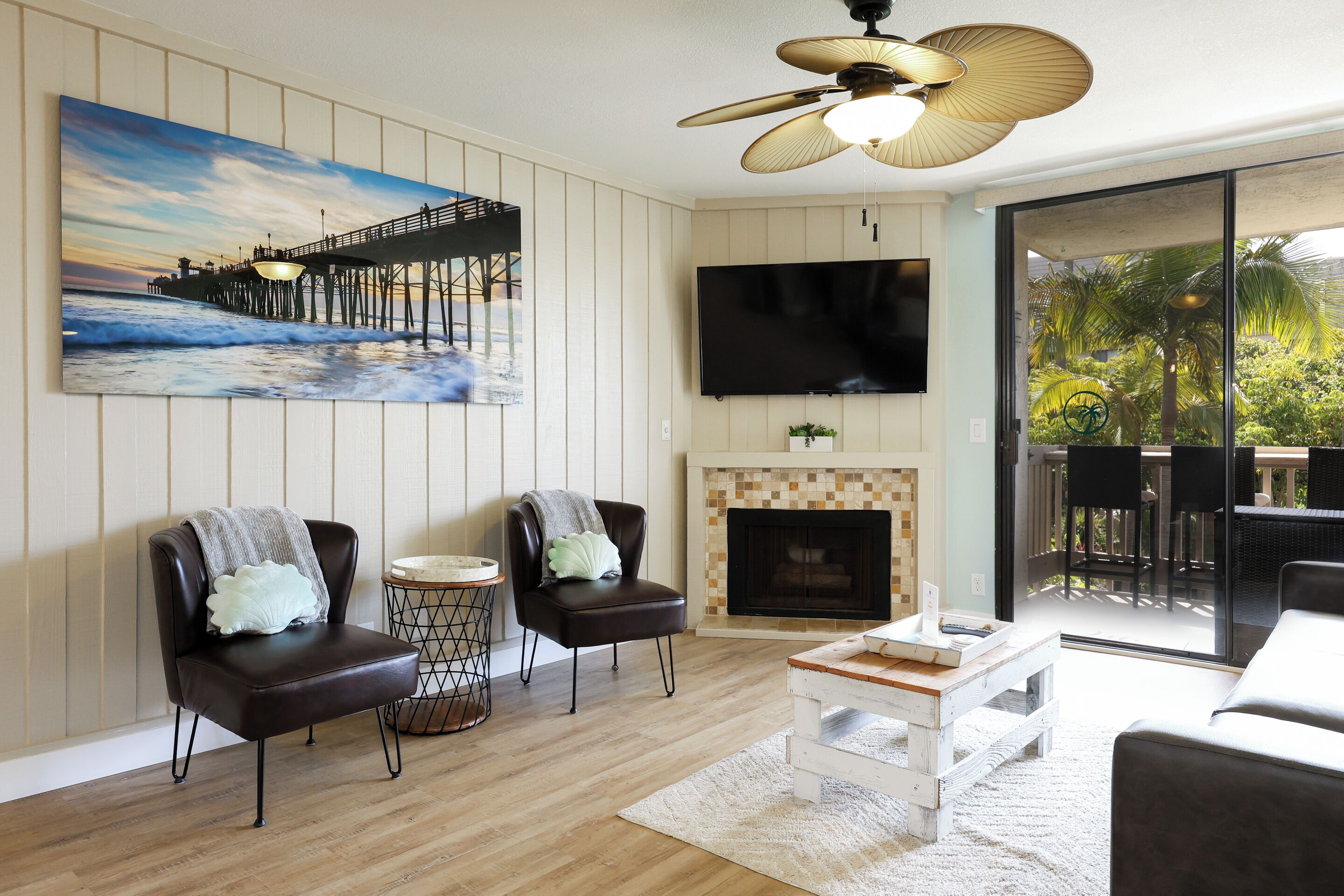 A living room with a wooden floor, two leather chairs, a white coffee table, a fireplace, a TV, and a large window with a view of palm trees.