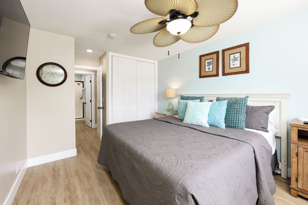 A bedroom with a bed, a ceiling fan, a clock, and a door leading to another room.