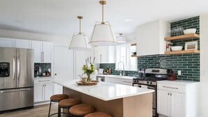 A modern kitchen with white cabinets, a central island with stools, stainless steel appliances, and green-tiled backsplash.