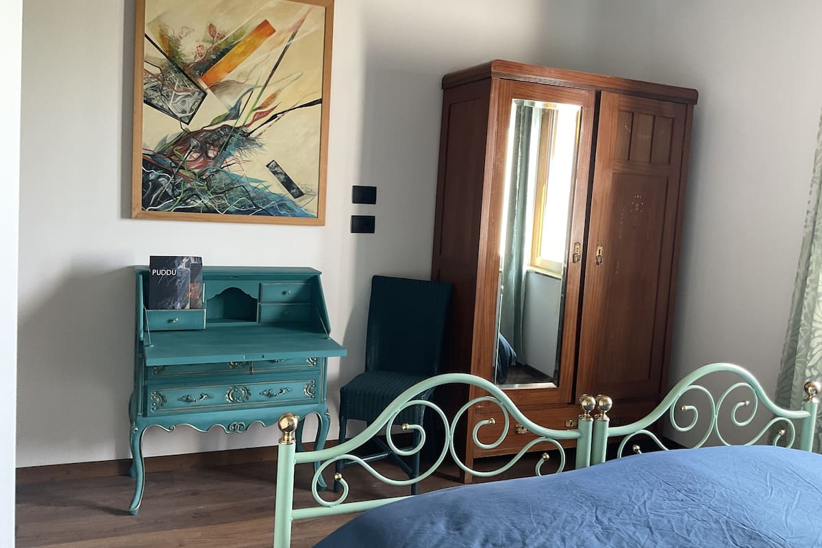 A bedroom with a bed, a teal dresser, a wooden wardrobe, and a painting on the wall.