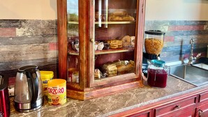 Une cuisine avec un meuble d’exposition en bois contenant du pain et des pâtisseries, une bouilloire, un bocal de céréales Nestlé Oorzo et une machine à café.