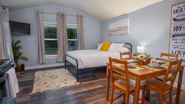 Ein Schlafzimmer mit Bett, Esstisch mit Stühlen, einem an der Wand befestigten Fernseher und einem Fenster mit Vorhängen.