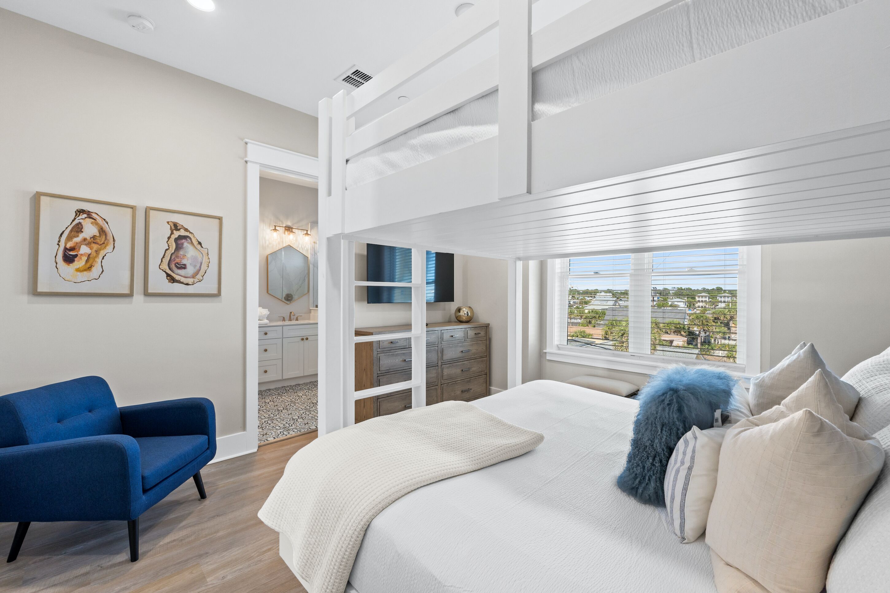 A bedroom with a bunk bed, a blue chair, a bedside table, and a view of the city through large windows.