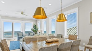 Un salón amplio con mesa de comedor de madera, sillas de mimbre y vista al océano a través de ventanas grandes.