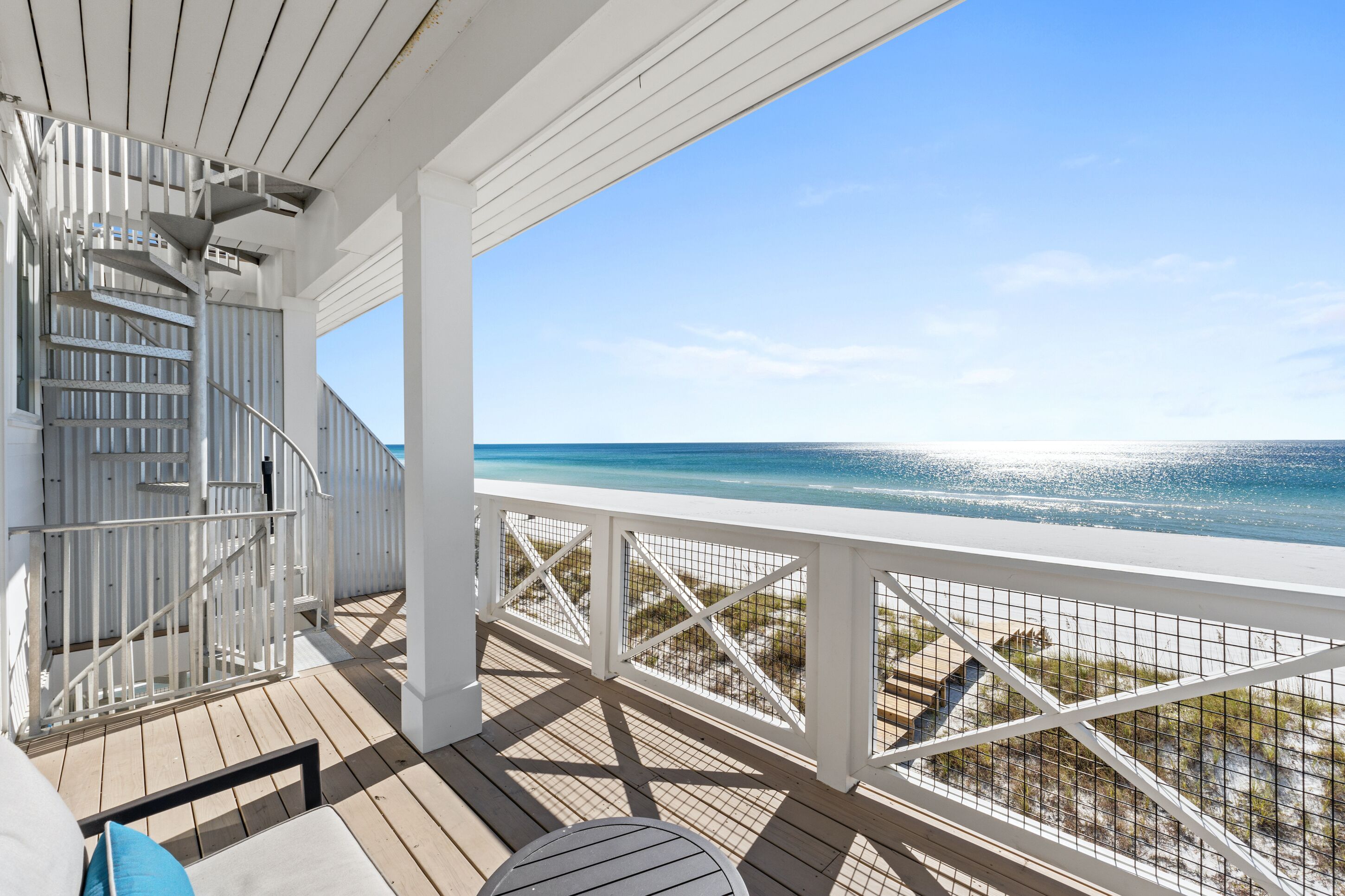Una terraza de madera con barandilla blanca, una escalera y vista a la playa.