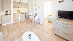 Eine moderne Küche mit weißen Schränken, eine Essecke mit weißem Tisch und Stühlen, ein Fernseher auf einem Schrank und ein kleiner Teppich auf dem Boden.