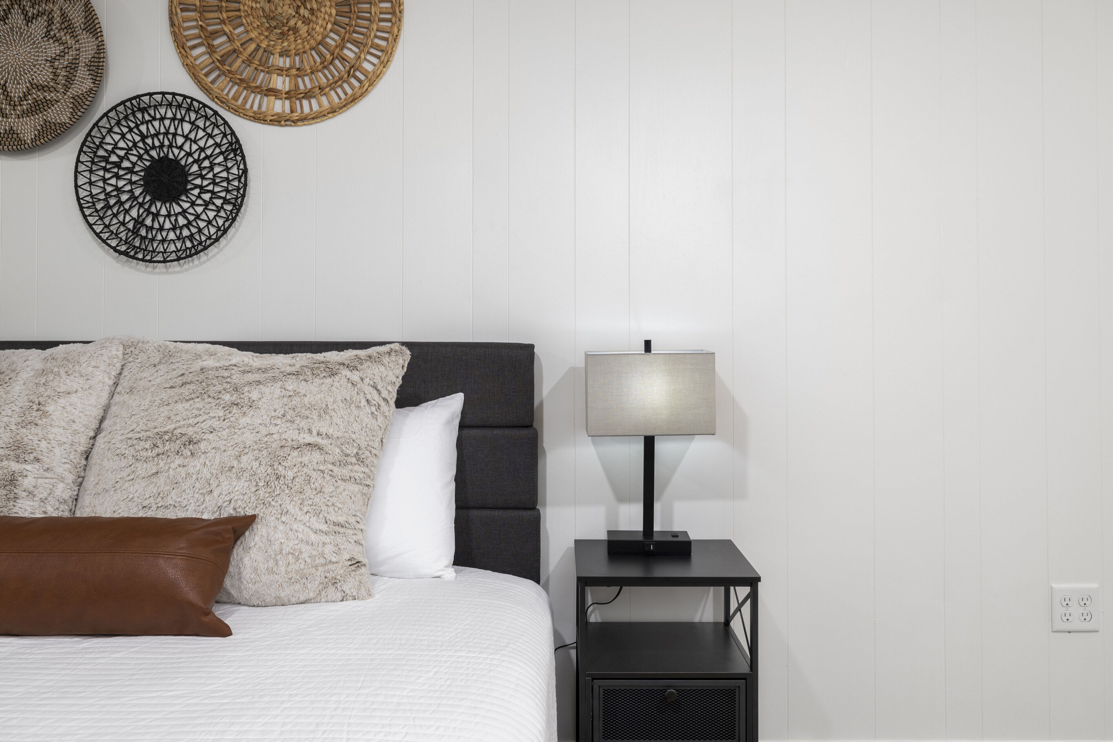 A neatly made bed with a white comforter, a brown pillow, and a grey headboard. A black nightstand with a lamp and a power outlet is beside the bed.