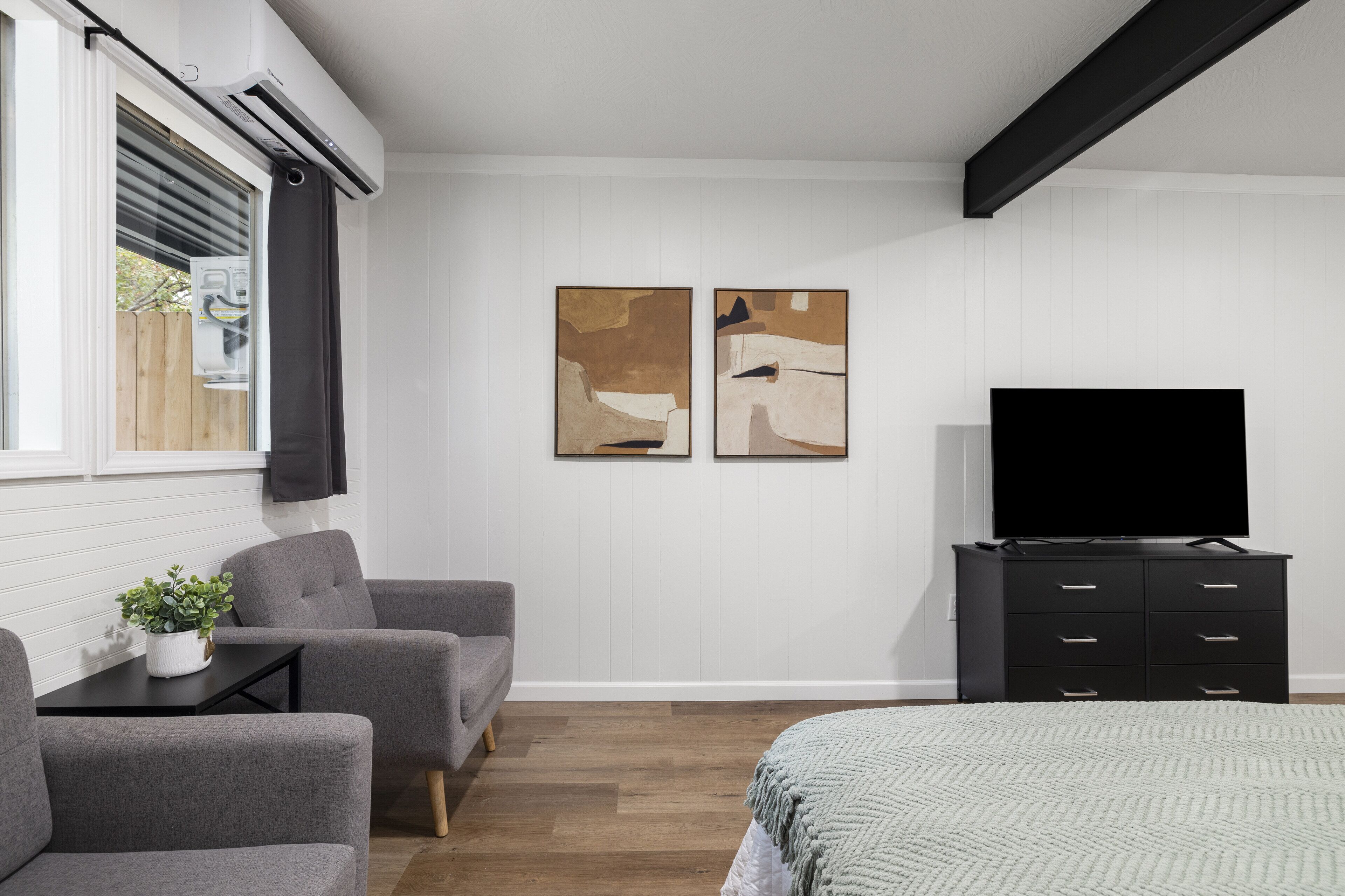 A modern room with a bed, two grey sofas, a TV on a black cabinet, and two abstract paintings on the wall.