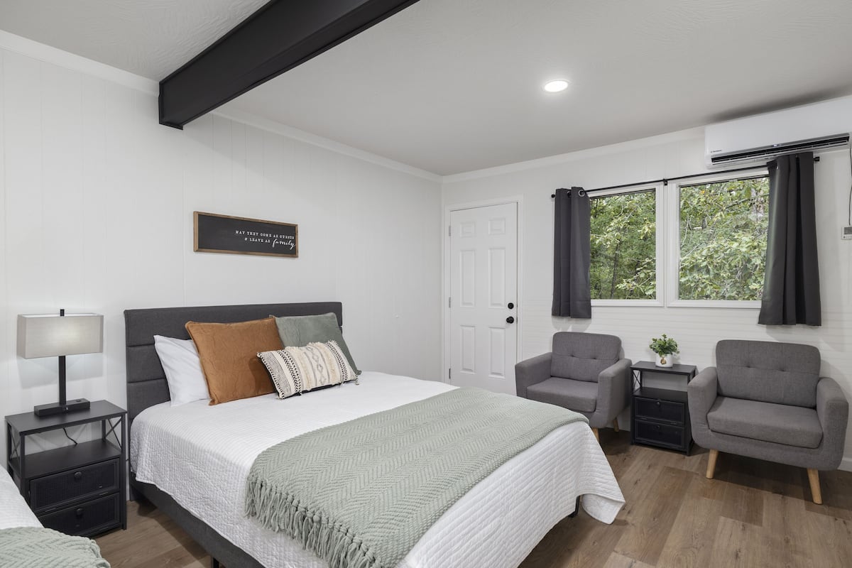 A modern bedroom with a large bed, two armchairs, a nightstand, and a window with curtains.