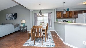 A modern kitchen with wooden cabinets, a dining area with a table and chairs, and a gray living room with a gray sofa. - Stoney Oasis (Charleston)