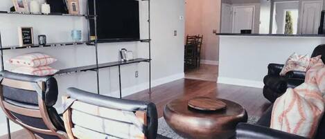Un salon moderne avec un canapé en cuir noir, une table basse en bois et un téléviseur fixé sur une étagère en métal noir. On aperçoit un coin repas à l’arrière-plan.