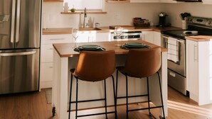 Una cocina moderna con armarios blancos, un refrigerador de acero inoxidable, un microondas y una encimera de madera. Hay dos taburetes y una ventana sobre el fregadero.