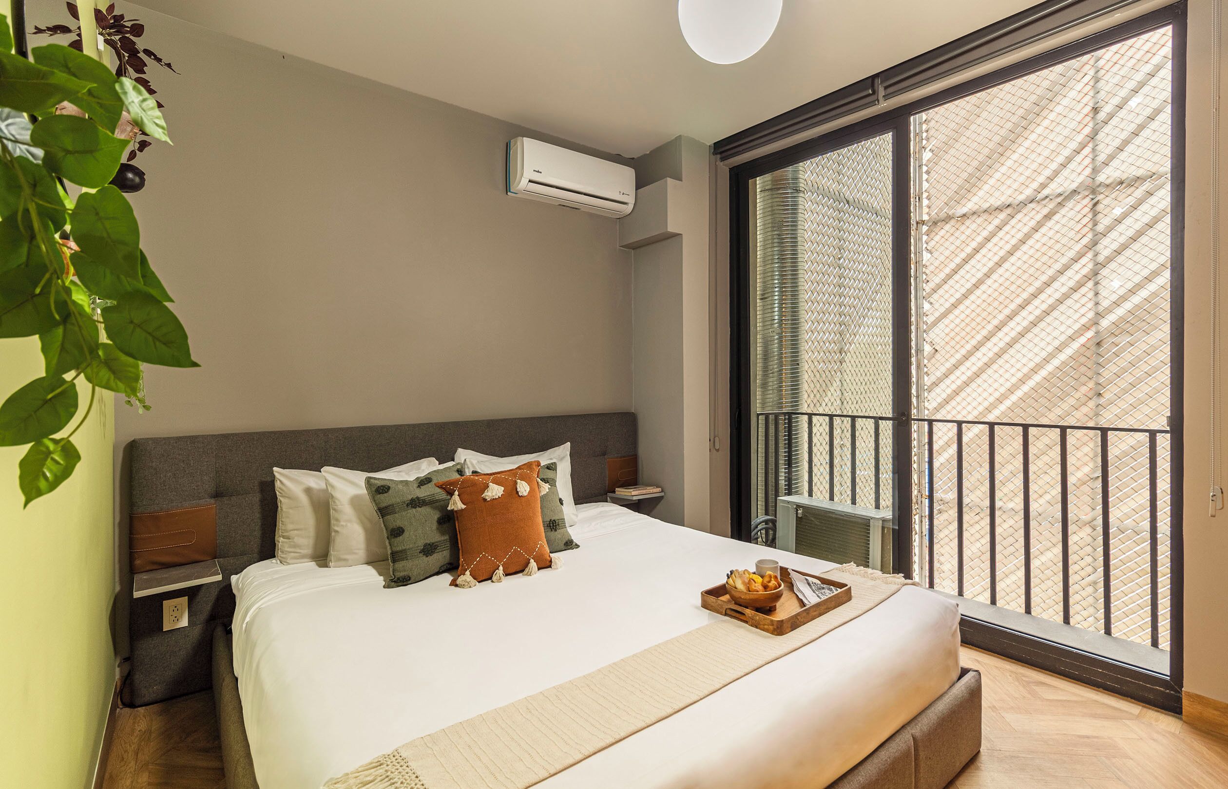 A modern bedroom with a large bed, a bedside table, an air conditioning unit, and a view through a large window.