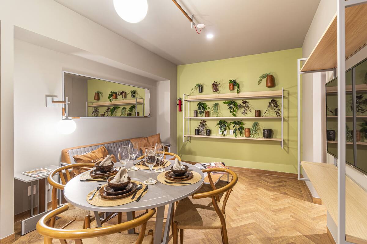 Un comedor con una mesa redonda dispuesta para cuatro personas, rodeada de sillas de madera. Hay varios estantes con macetas en las paredes.