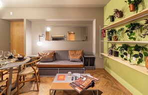 Ein modernes Wohnzimmer mit einer Couch, einem Esstisch mit Stühlen, einer Zeitschrift und einem Couchtisch. An der Wand befinden sich Regale mit Pflanzen.