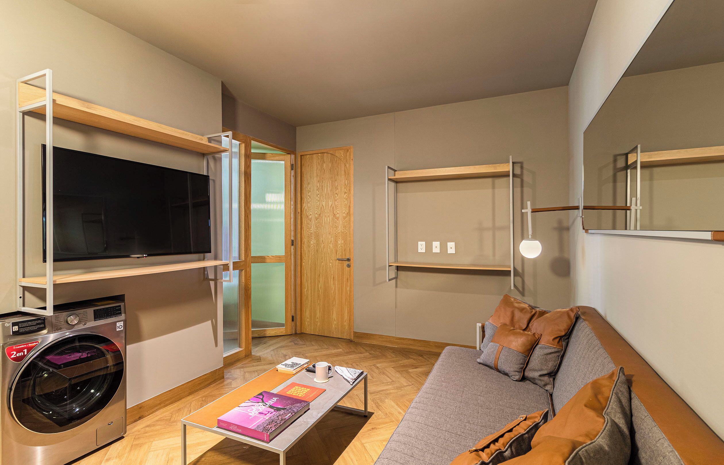 A compact living space with a flat-screen TV, a washing machine, a sofa, and a small table with books and a cup.