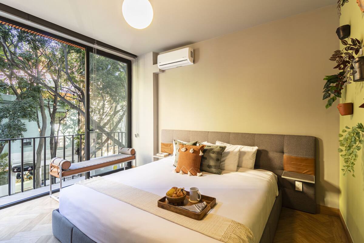 Habitación de hotel moderna con una cama grande, un balcón con vistas y una mesa pequeña con alimentos para el desayuno.