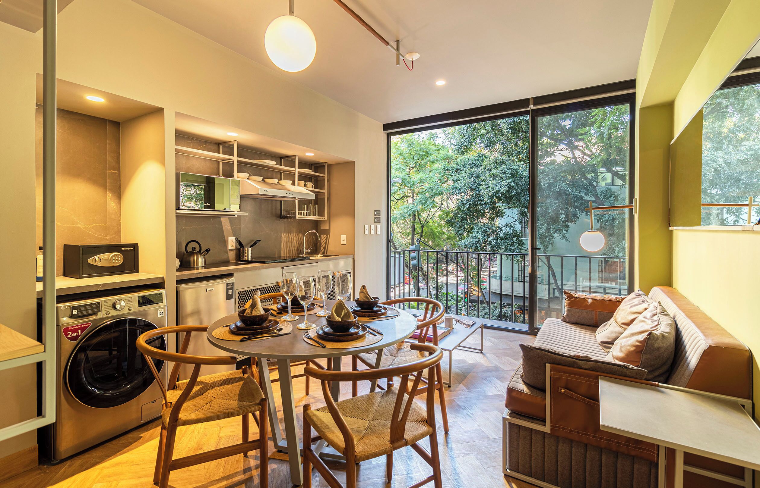 A modern kitchen with built-in appliances, a dining area with a round table and chairs, and a balcony with a view of trees.