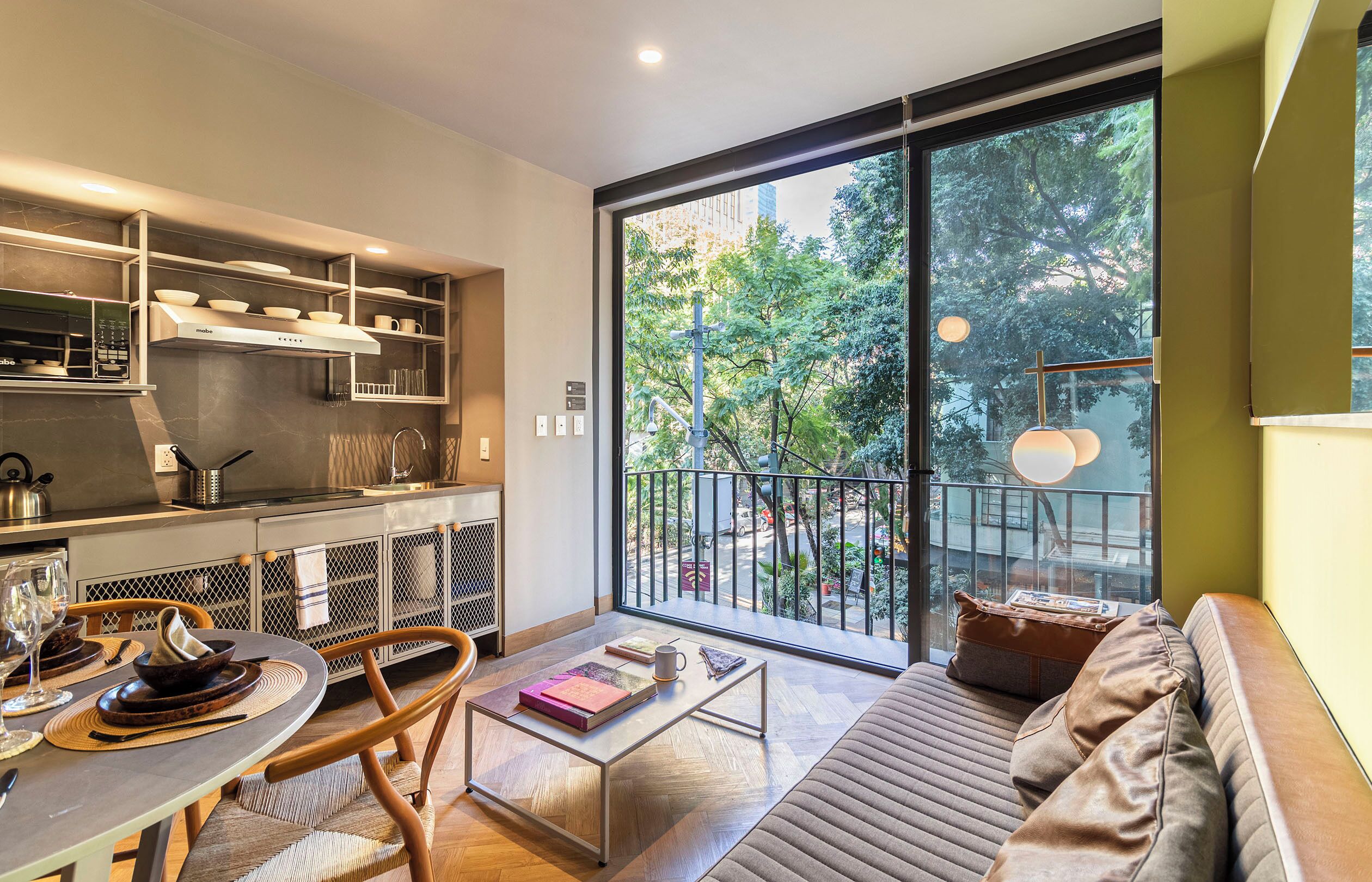 A modern kitchen with built-in appliances, a dining area with a round table and chairs, and a large sliding glass door leading to a balcony with a view of trees and a street.