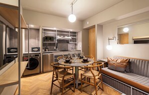 Eine kompakte Küche mit modernen Geräten, ein Essbereich mit rundem Tisch und Stühlen sowie eine Sitzecke mit Sofa und Kissen.