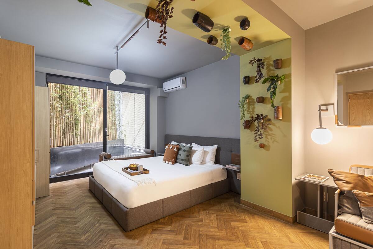 Un dormitorio moderno con una cama grande, una mesita de noche pequeña, una mesita auxiliar, un aire acondicionado de pared y una ventana con vistas a bambú.