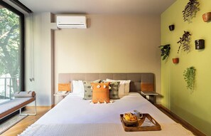 Une chambre d’hôtel avec un grand lit, des tables de chevet, un banc, un plateau avec des articles pour le petit-déjeuner et un climatiseur fixé au mur.